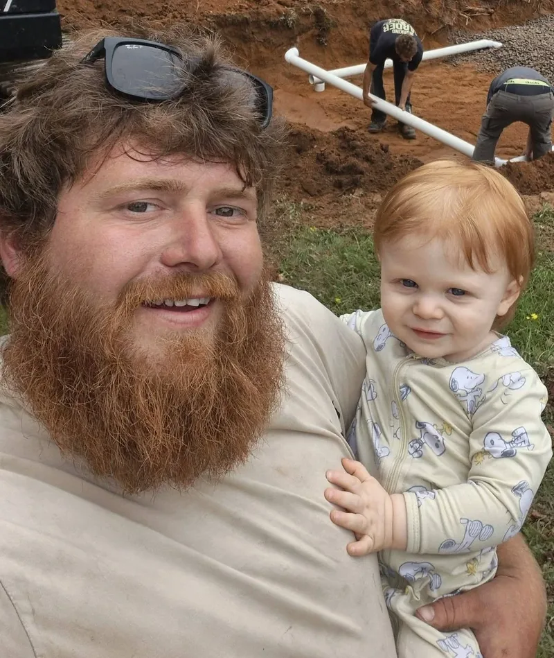 Brandon Aldred, owner of Aldred Excavation, holding his young child on a job site while his crew installs a septic system behind them